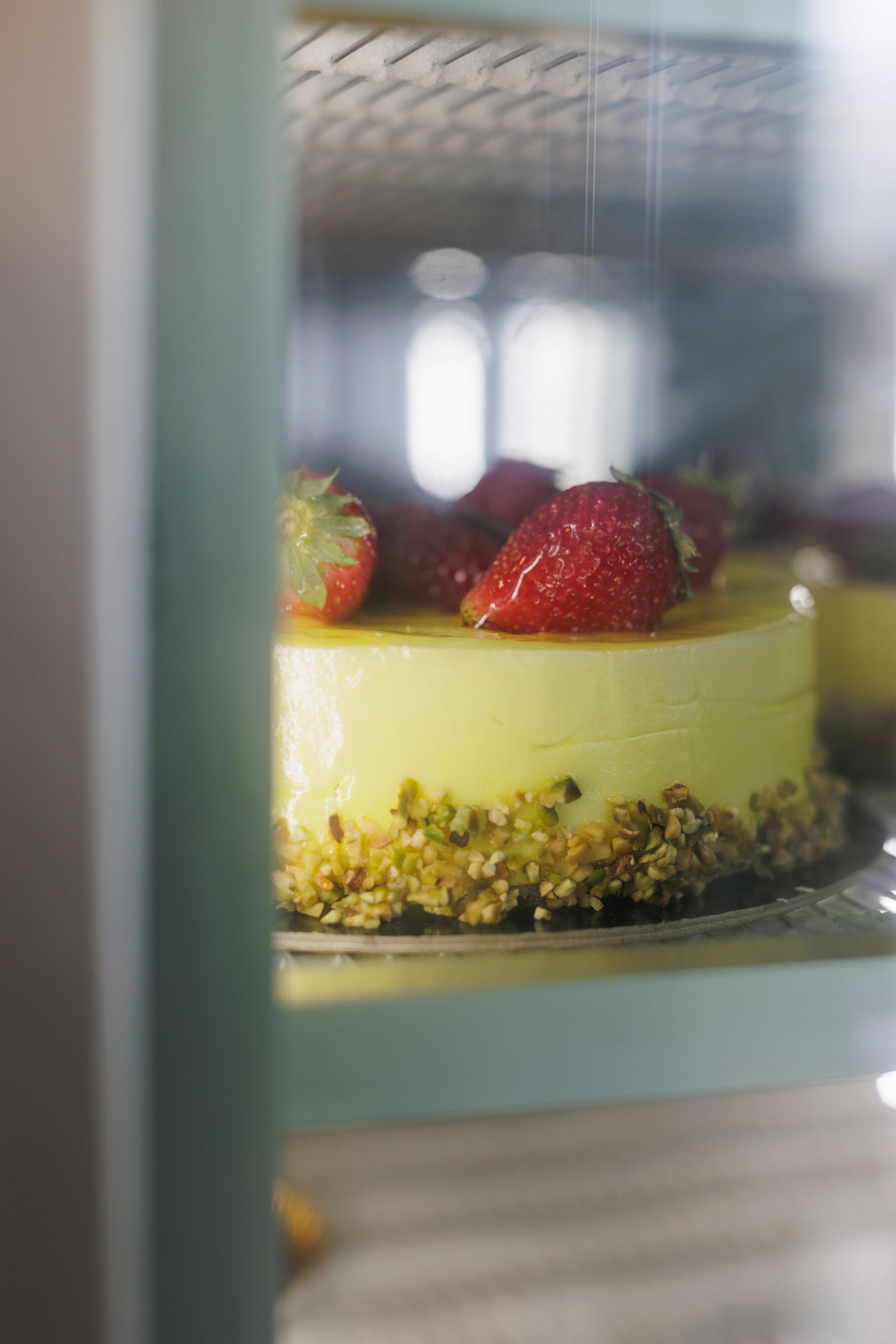 Una torta gialla e soffice, guarnita con fragole fresche e una crosta di pistacchi tritati, vista attraverso una vetrina.