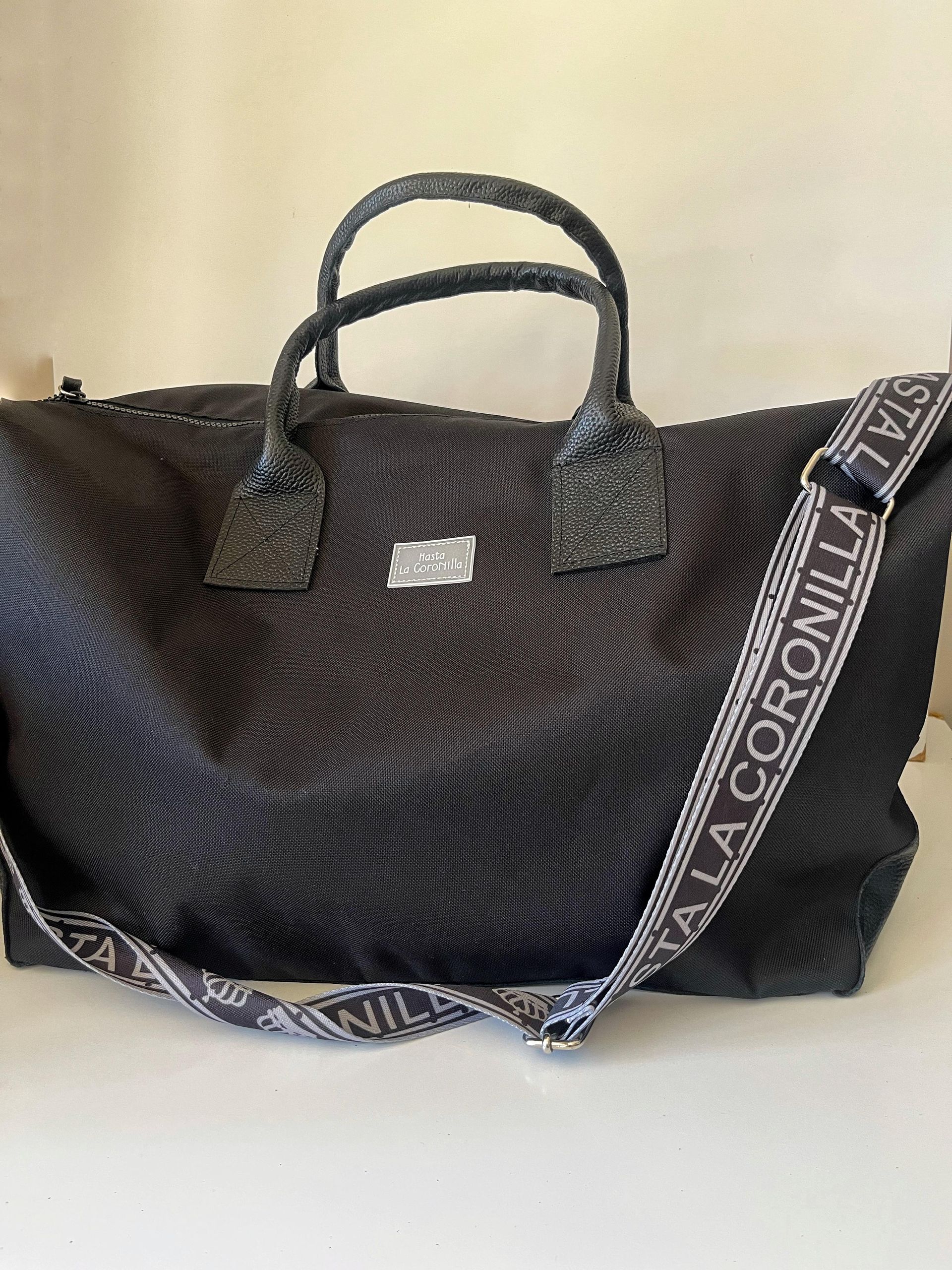Bolso de lona negro texturizado con dos asas superiores y una correa de hombro ancha con la marca 