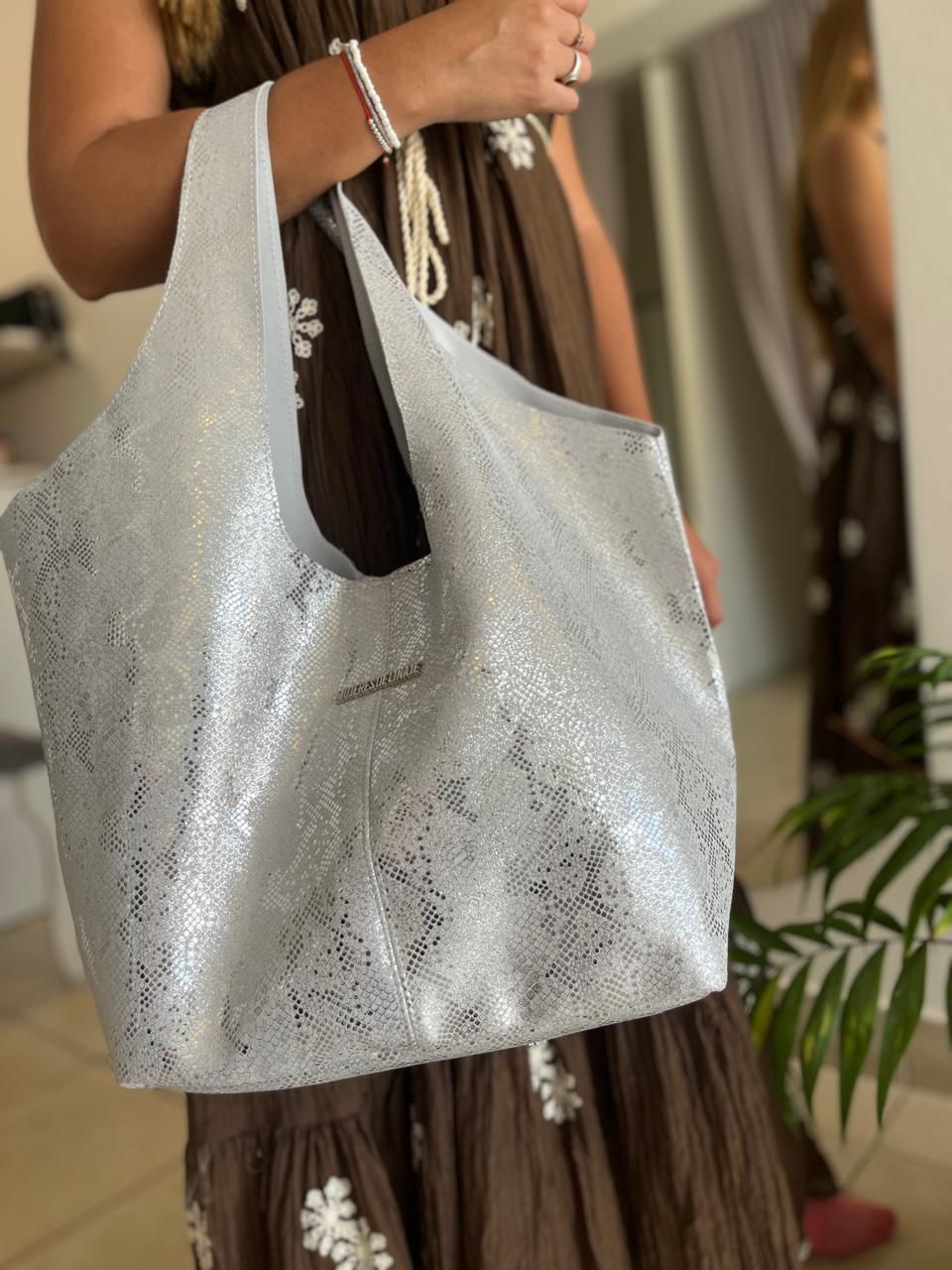 Un bolso de mano plateado y texturizado sostenido por una persona que viste un vestido marrón con bordados florales blancos.