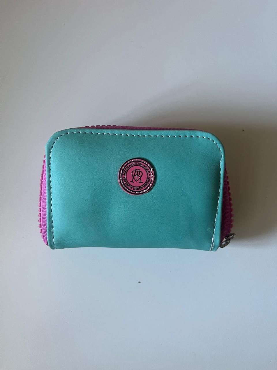 Un monedero pequeño, rectangular, de color verde azulado, con una cremallera rosa