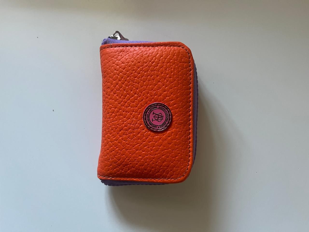 Un monedero pequeño, de piel granulada, de color naranja, con cremallera de color violeta y un emblema circular en el centro.