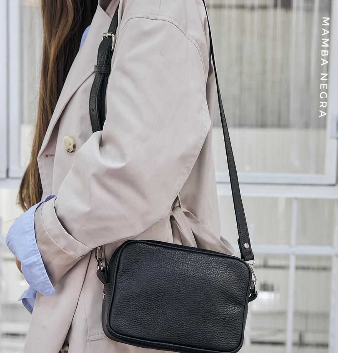 Una persona con una gabardina beige y una camisa azul claro lleva un bolso bandolera de cuero texturizado de color negro.