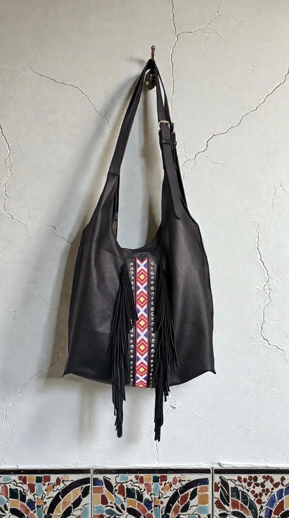 Un bolso hobo de cuero oscuro cuelga de una pared y presenta una banda central decorativa con flecos