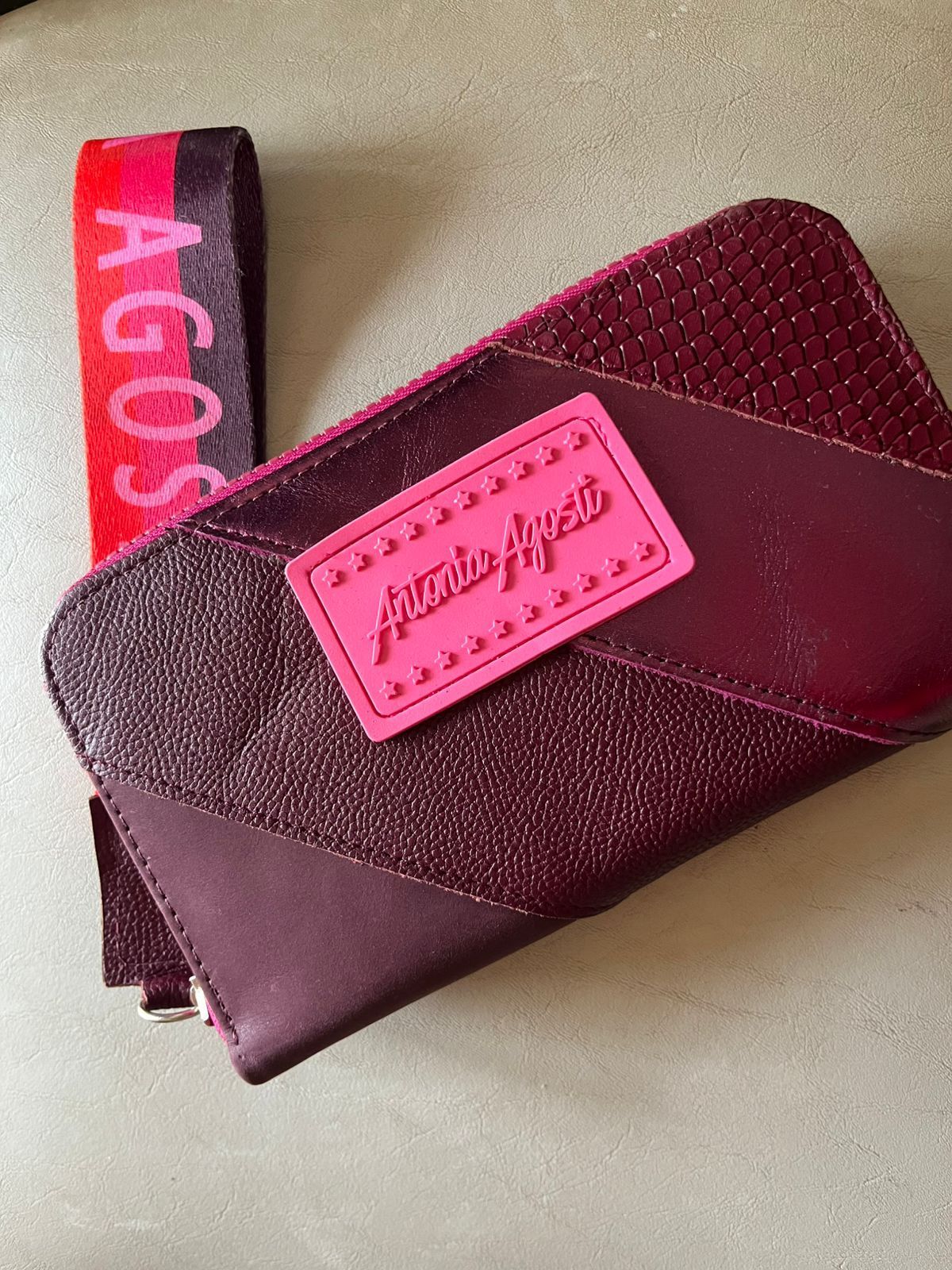 Una cartera de mano con textura de color granate con un parche de logotipo rosa y una correa de muñeca de marca