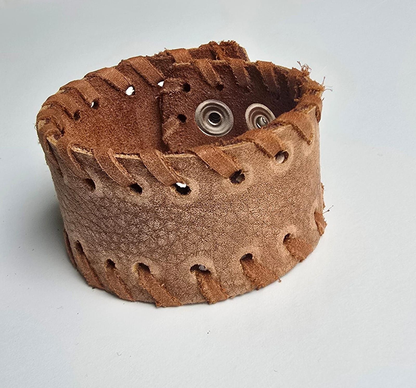 Una pulsera ancha de cuero marrón con cordones decorativos en los bordes y dos cierres a presión de metal.