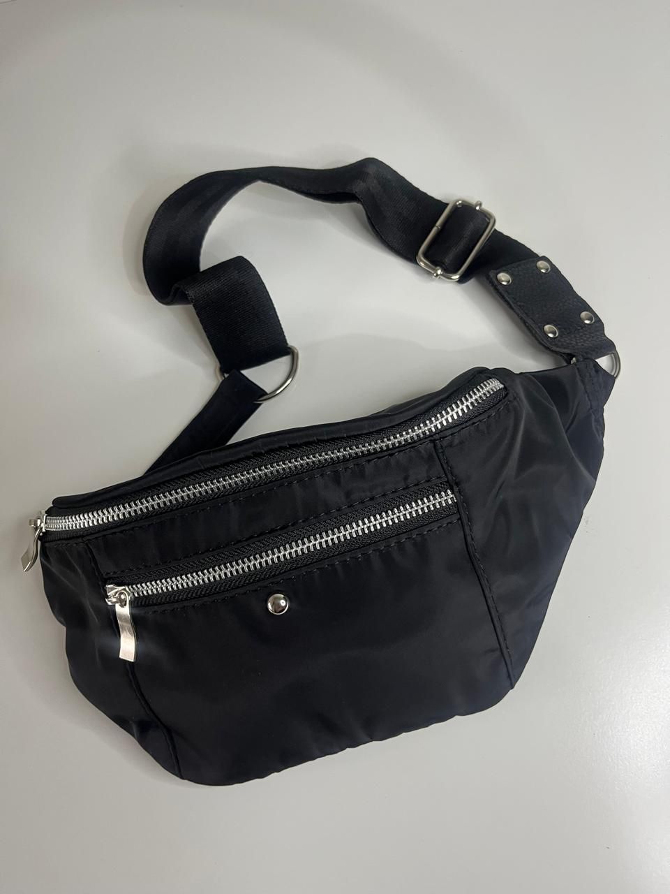 Un bolso de cintura negro con cremalleras plateadas y una correa ajustable, centrado sobre un fondo blanco liso.