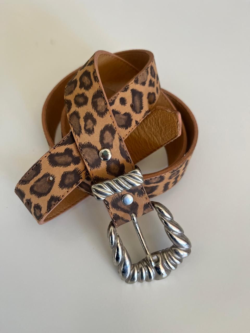 Un cinturón con estampado de leopardo enrollado sobre una superficie blanca con una hebilla de metal retorcido plateado.