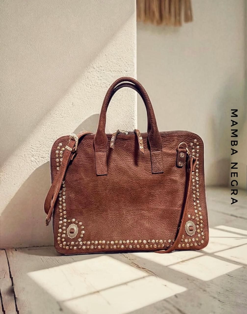 Un bolso de gamuza marrón con bordes tachonados, detalles y asas plateadas, sobre una superficie de color claro.