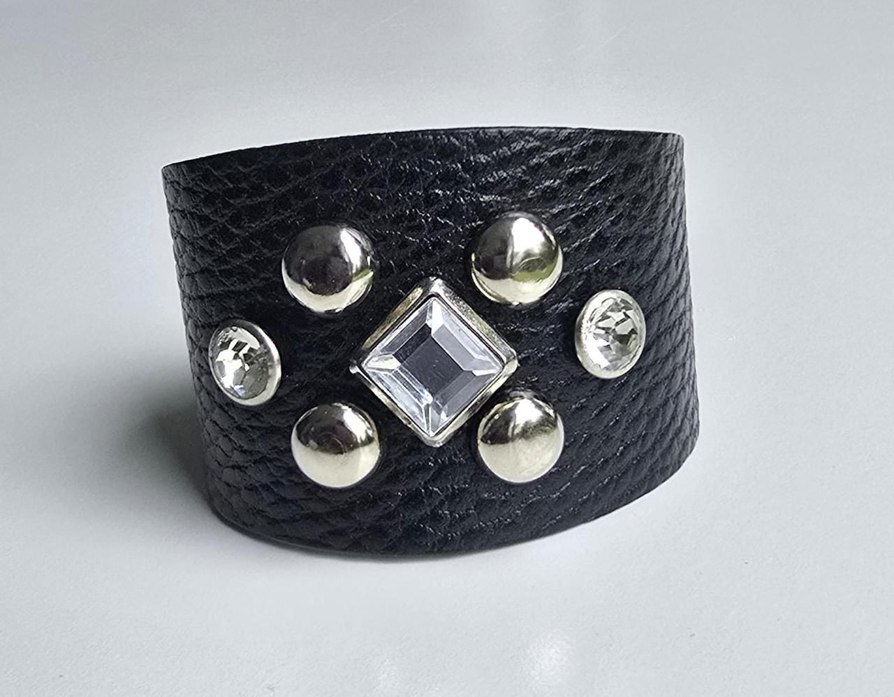 Una pulsera de cuero negro que presenta un cristal cuadrado transparente central acentuado por cuatro tachuelas plateadas