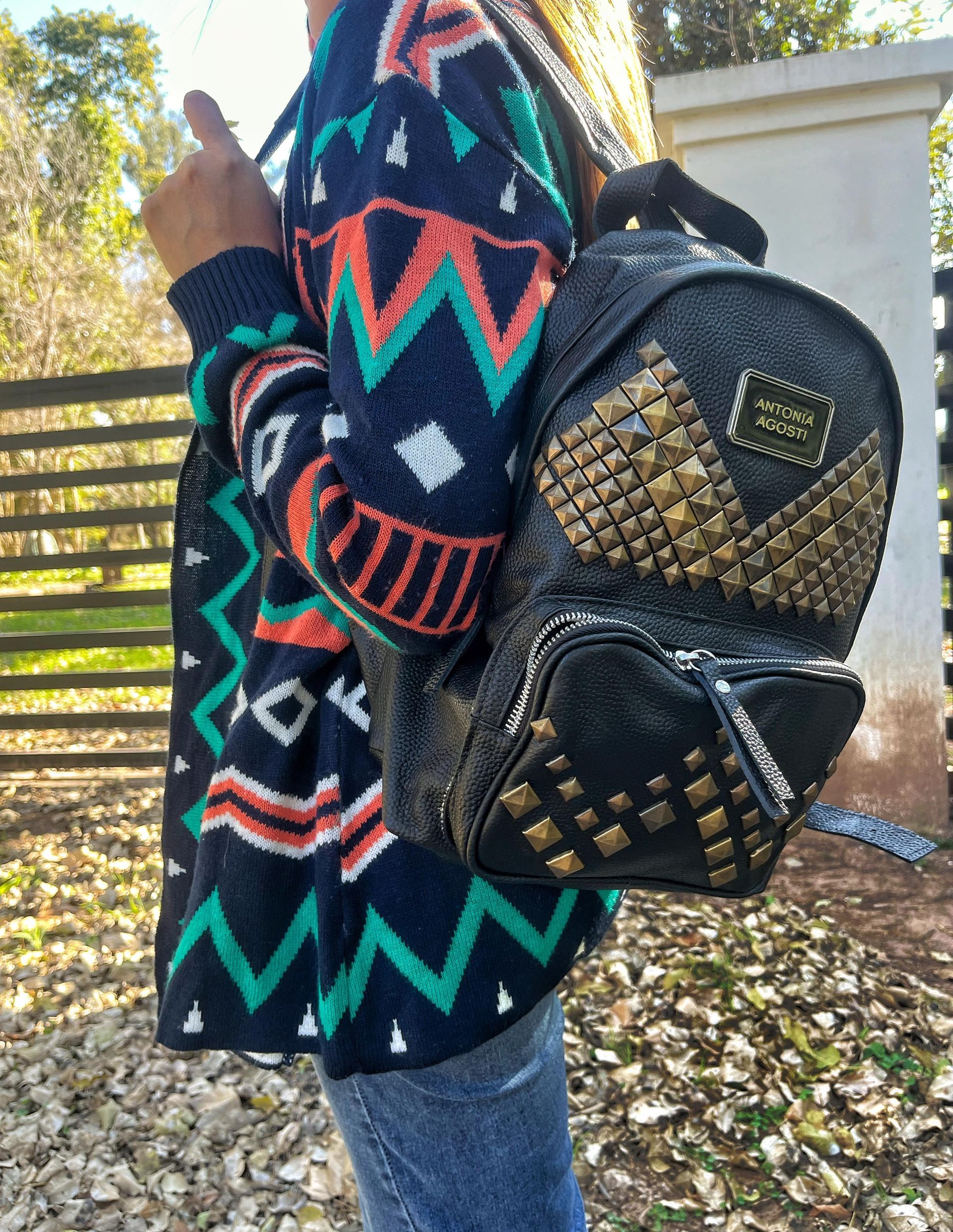 Una persona viste un cárdigan colorido con estampado en zigzag y lleva una mini mochila negra con tachuelas al aire libre.