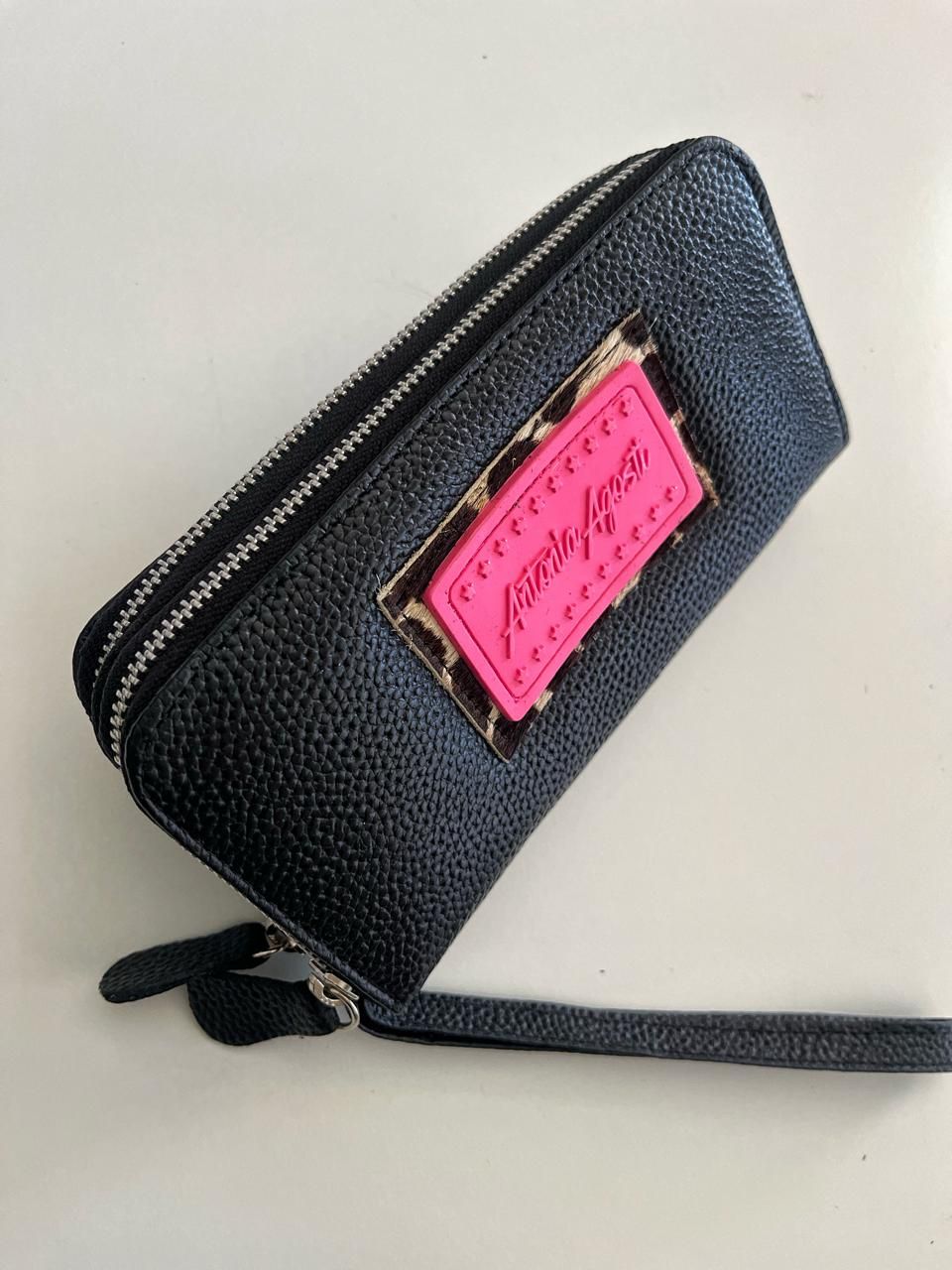 Una billetera de muñeca negra y texturizada con una placa de logotipo rectangular de color rosa neón