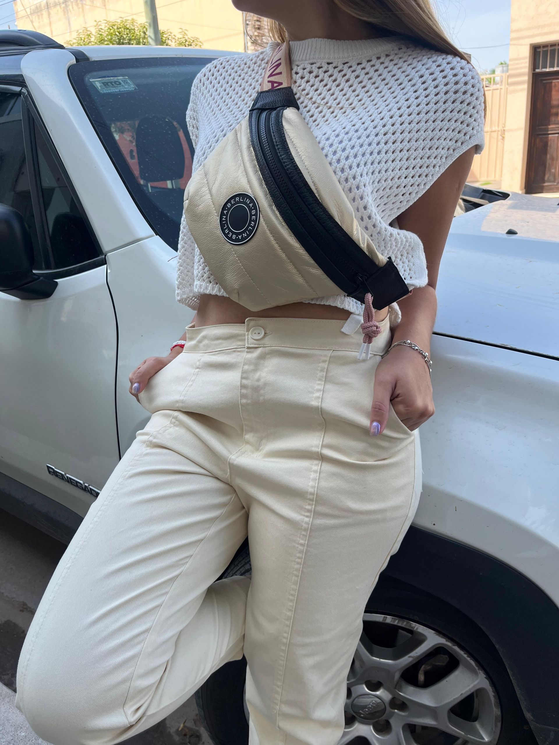 Una persona está parada al lado de un vehículo blanco, vistiendo una blusa de punto blanca, pantalones de color crema