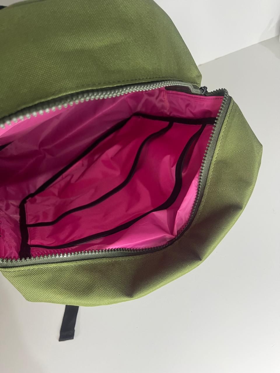 Mochila verde oliva con base negra, bolsillo frontal con cremallera y pestaña con forma de diamante negro sobre fondo blanco.
