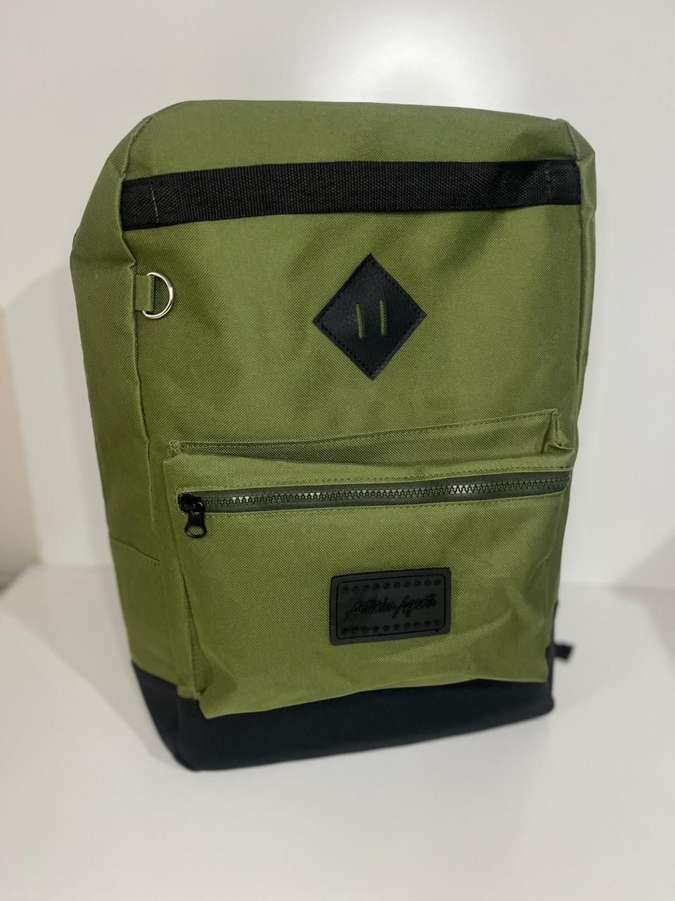Mochila verde oliva con base negra, bolsillo frontal con cremallera y pestaña con forma de diamante negro sobre fondo blanco.