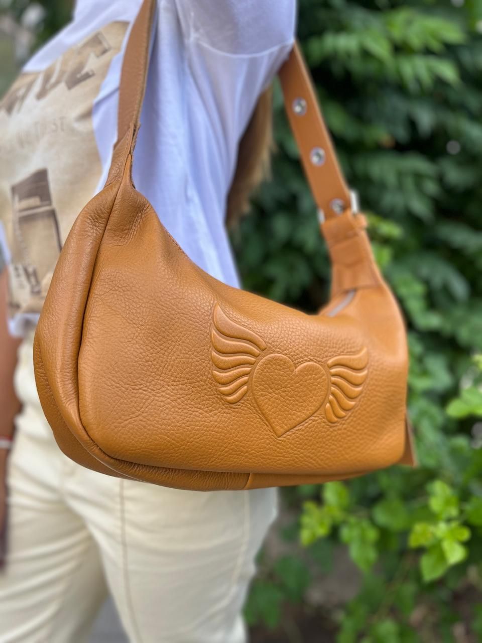 Un bolso estilo hobo de cuero color canela con un logotipo de corazón alado en relieve, que se usa sobre el hombro