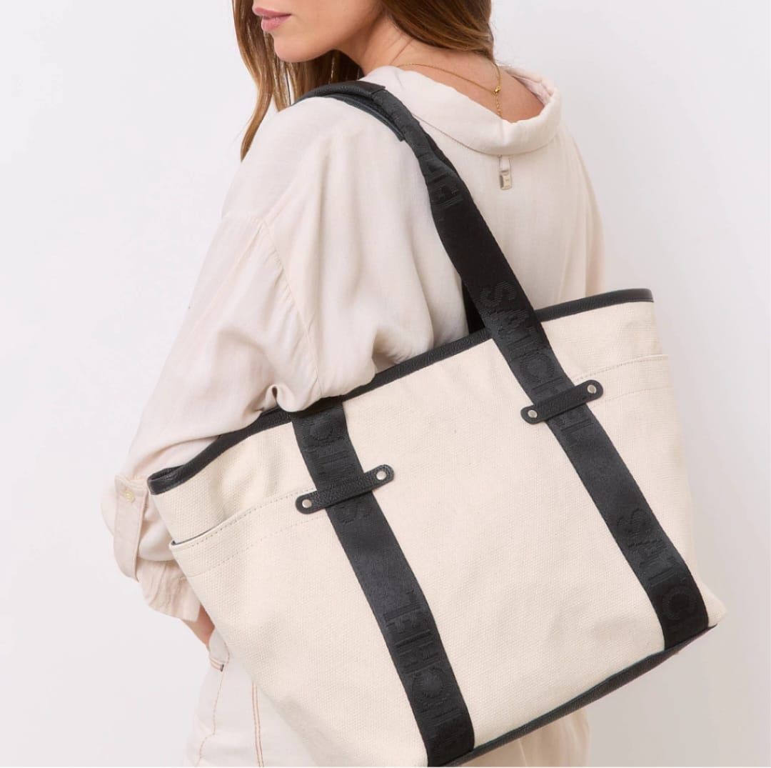 Una persona lleva un bolso de lona beige con correas negras sobre el hombro sobre un fondo blanco.