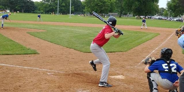 A person wearing a hat and orange shirt, holding a baseball bat on their shoulder on a baseball field.