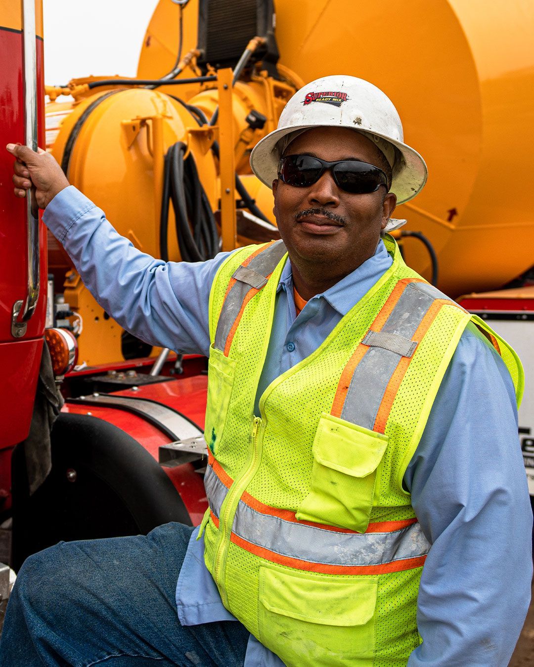 A man wearing a hard hat and sunglasses is sitting in front of a yellow truck.