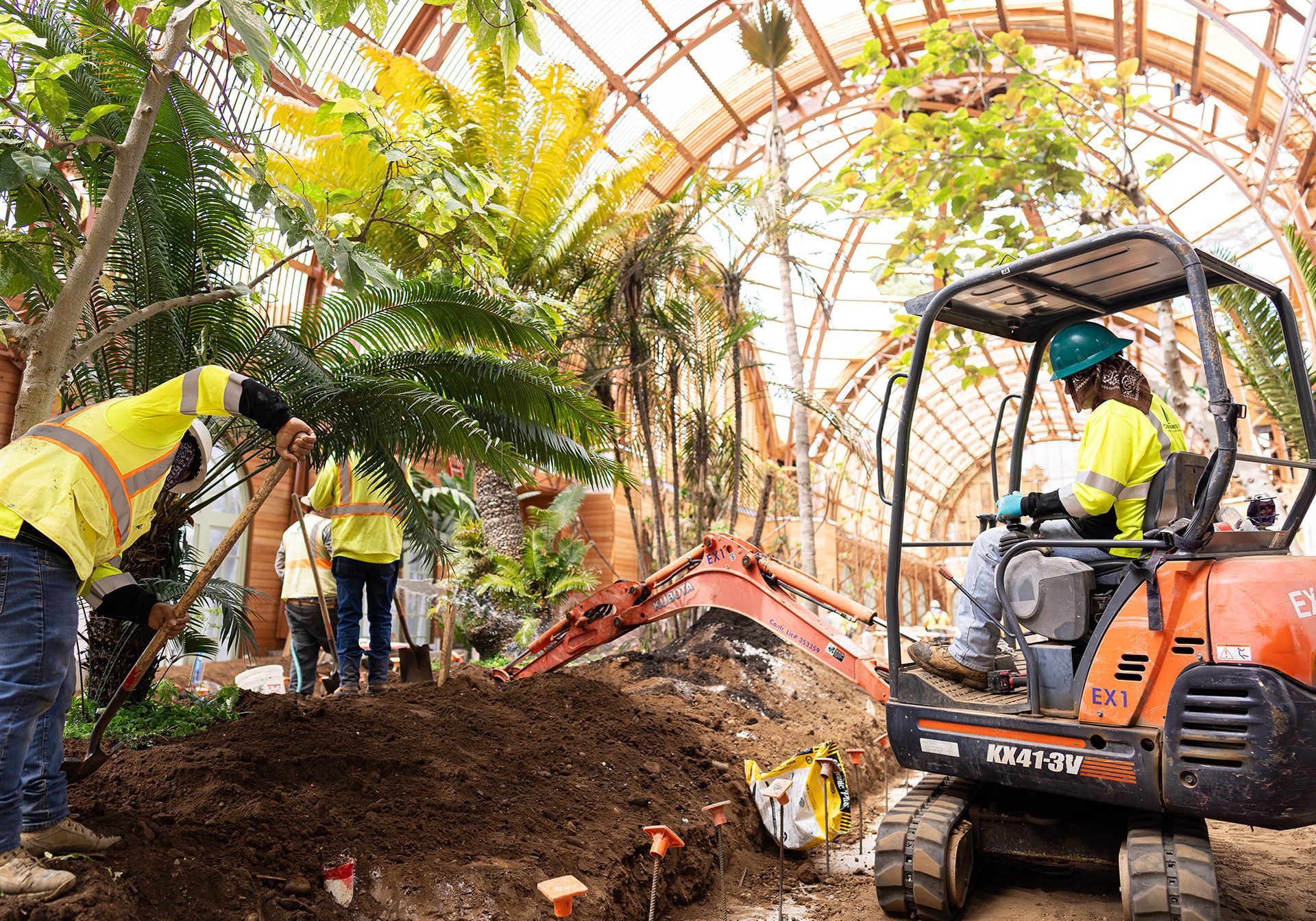 Workers in high-visibility vests operate a small orange excavator inside a large, glass-roofed greenhouse.