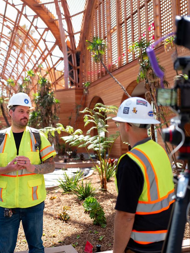 Two construction workers in vests and hard hats stand in a wooden structure with plants.