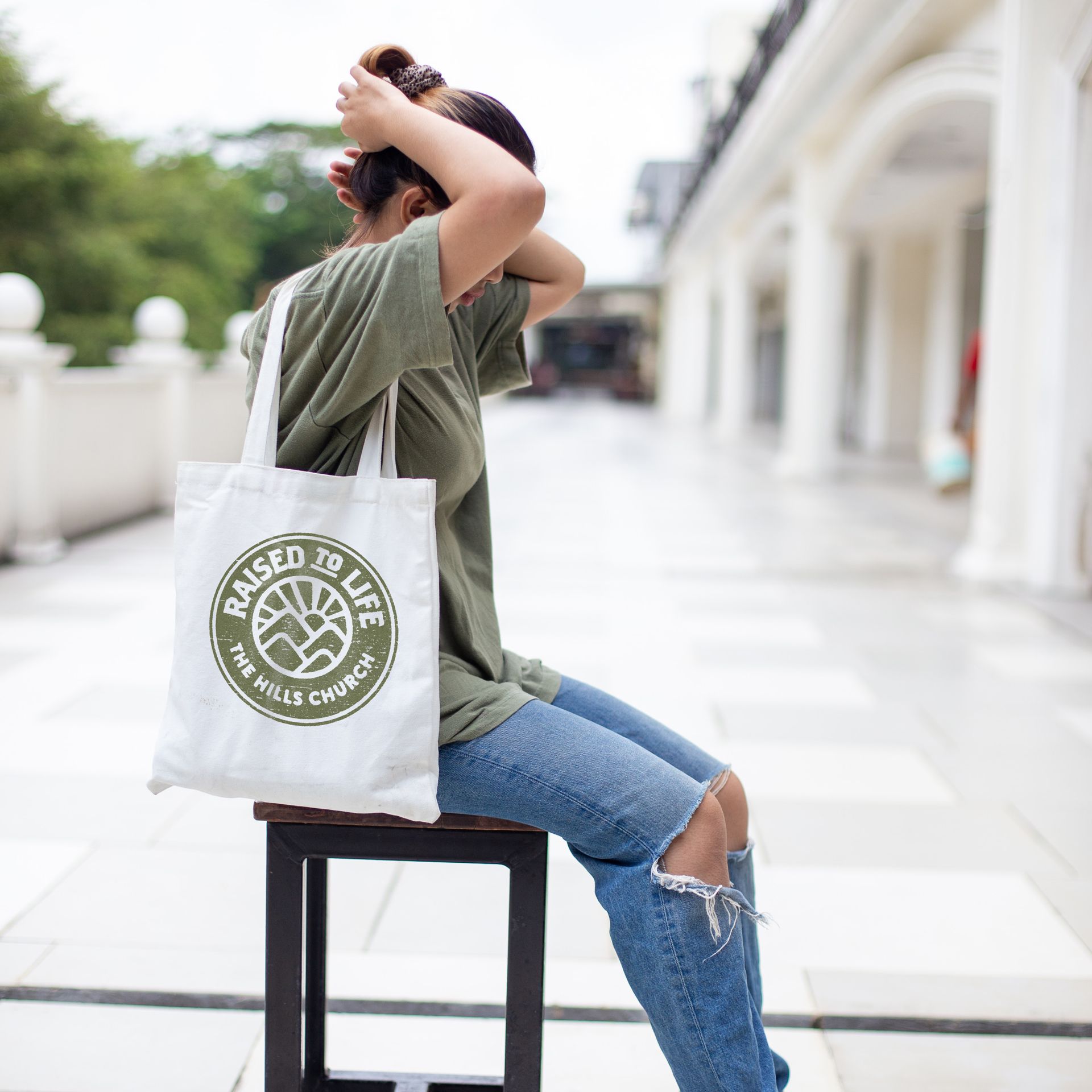 A woman is sitting on a stool holding a tote bag that says raised 29 life