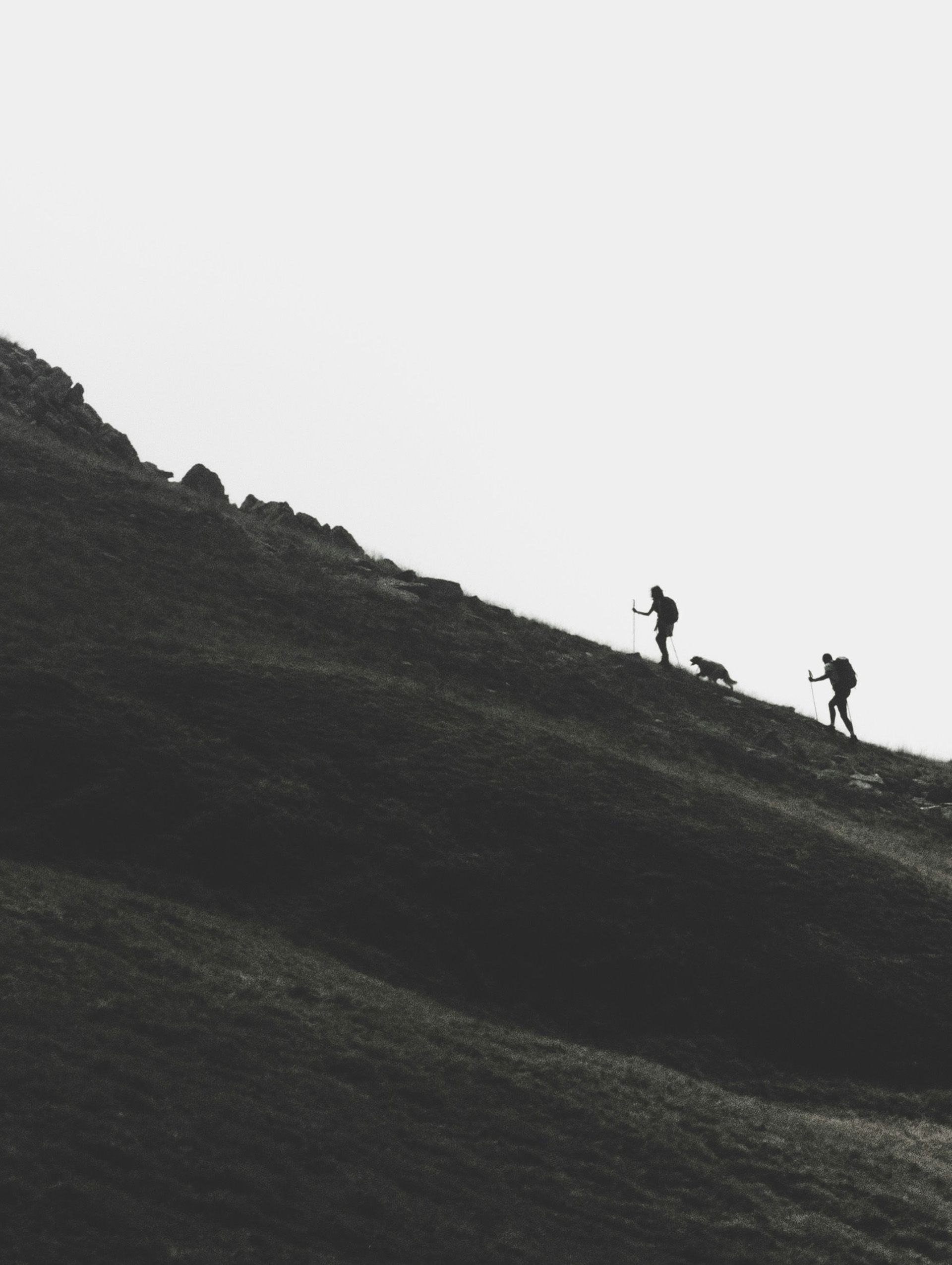 Hikers climb a steep, grassy mountain in monochrome.