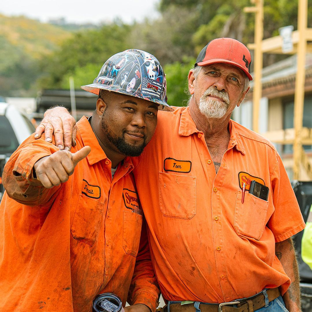 Two people in orange uniforms posing for a photo outdoors, one wearing a patterned hard hat and giving a thumbs up.