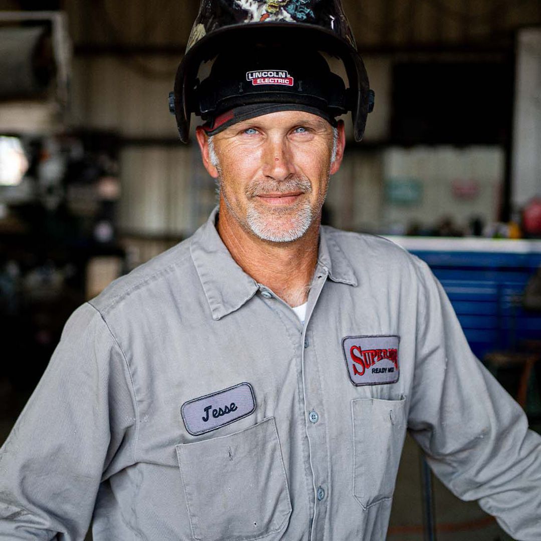 A person in a gray work shirt and a welding helmet worn on their head, standing in a workshop setting.