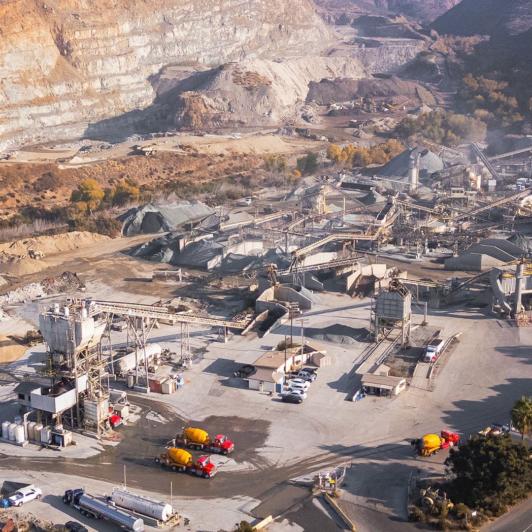 An aerial view of an industrial quarry featuring machinery, concrete mixers, piles of crushed rock, and rock-faced slopes.
