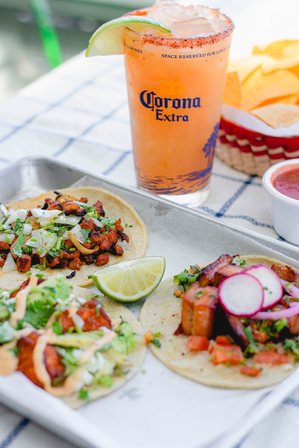 A tray of tacos and a glass of corona extra on a table.