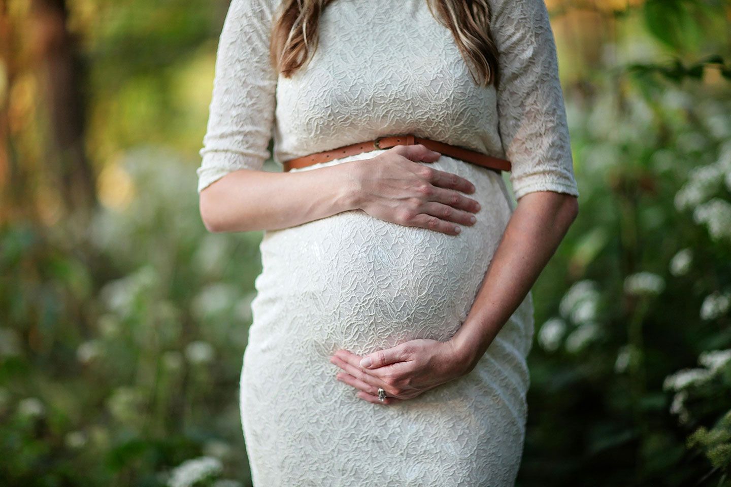 Pregnant woman in a white dress, hands cradling her belly, standing outdoors in front of greenery.