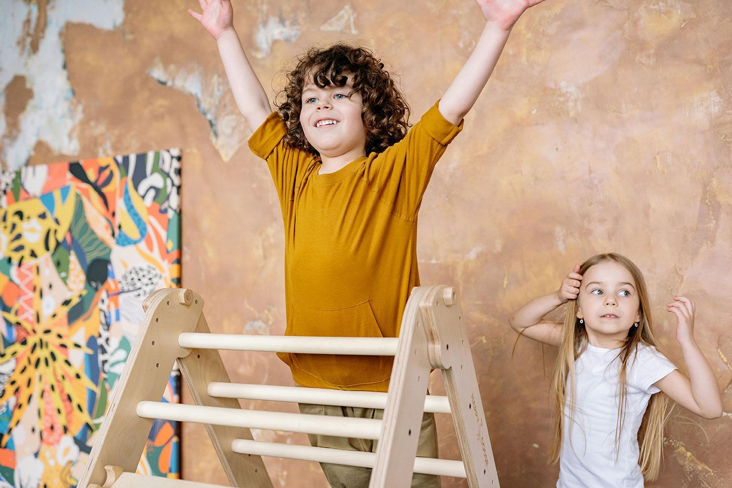 Boy with arms raised atop a climbing frame, girl watching with surprise, inside.