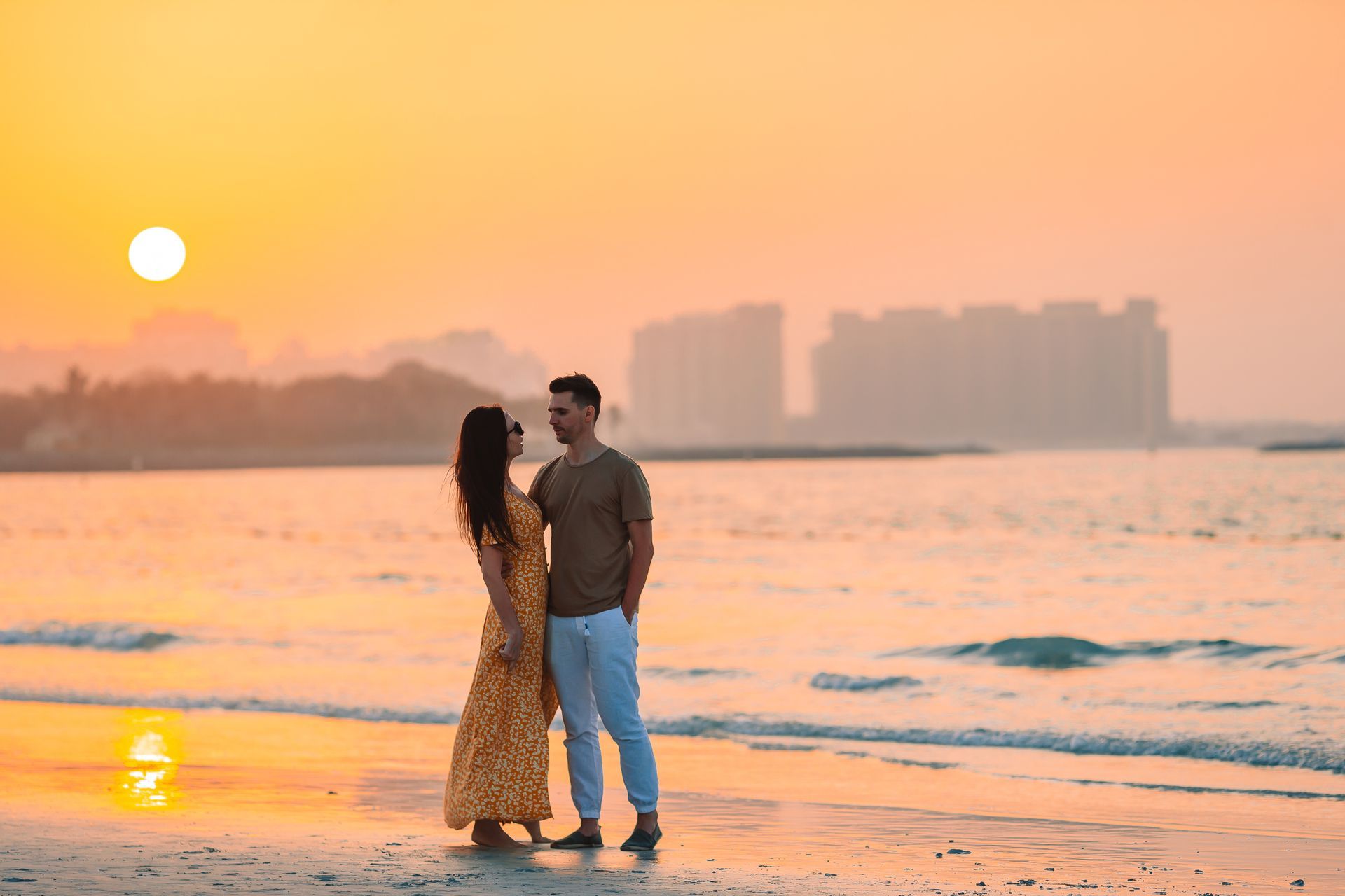 Couple standing on a beach at sunset with gentle waves and city skyline in the background