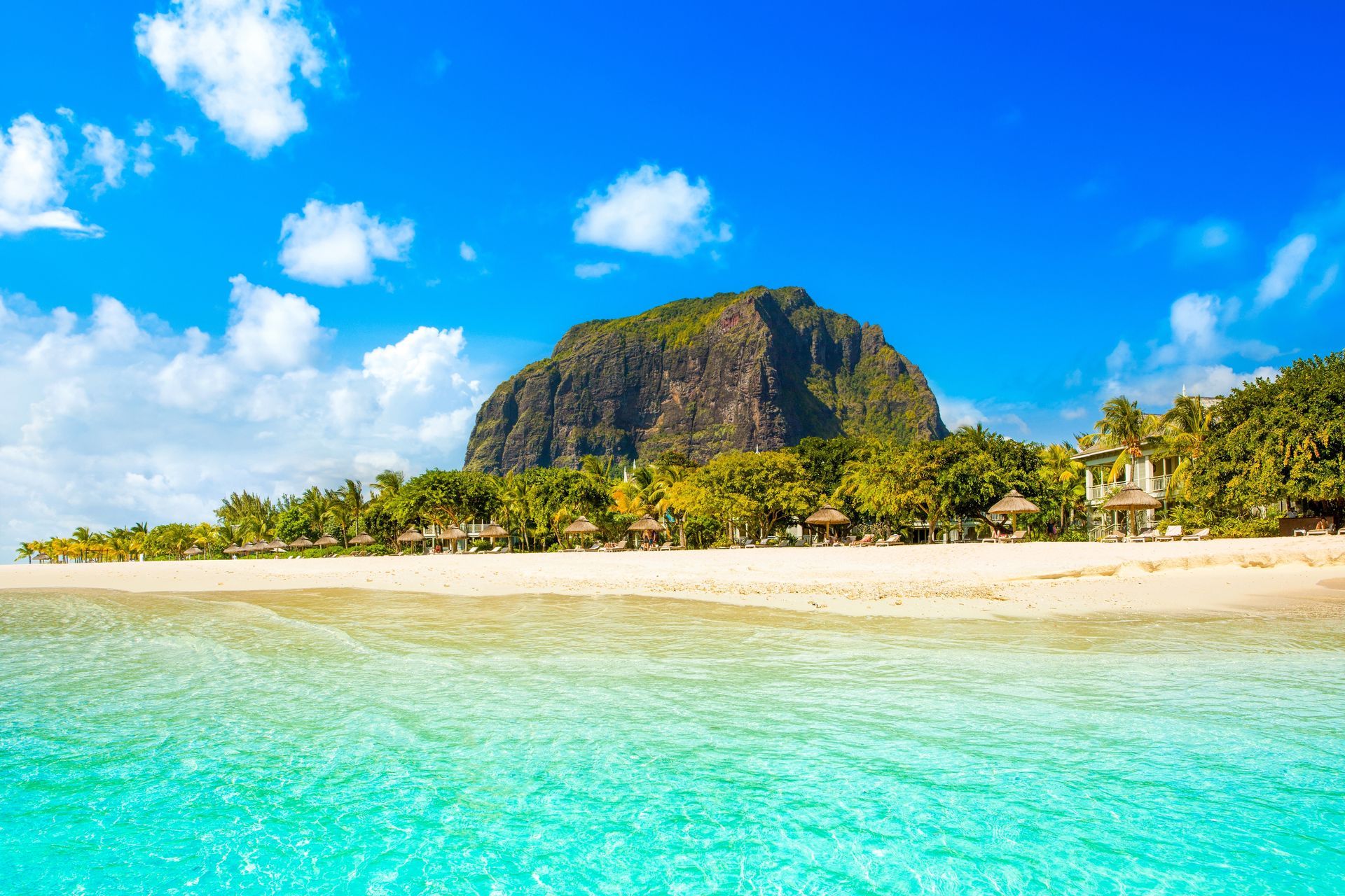 Tropical beach with turquoise water, white sand, and a large rocky hill under a bright blue sky