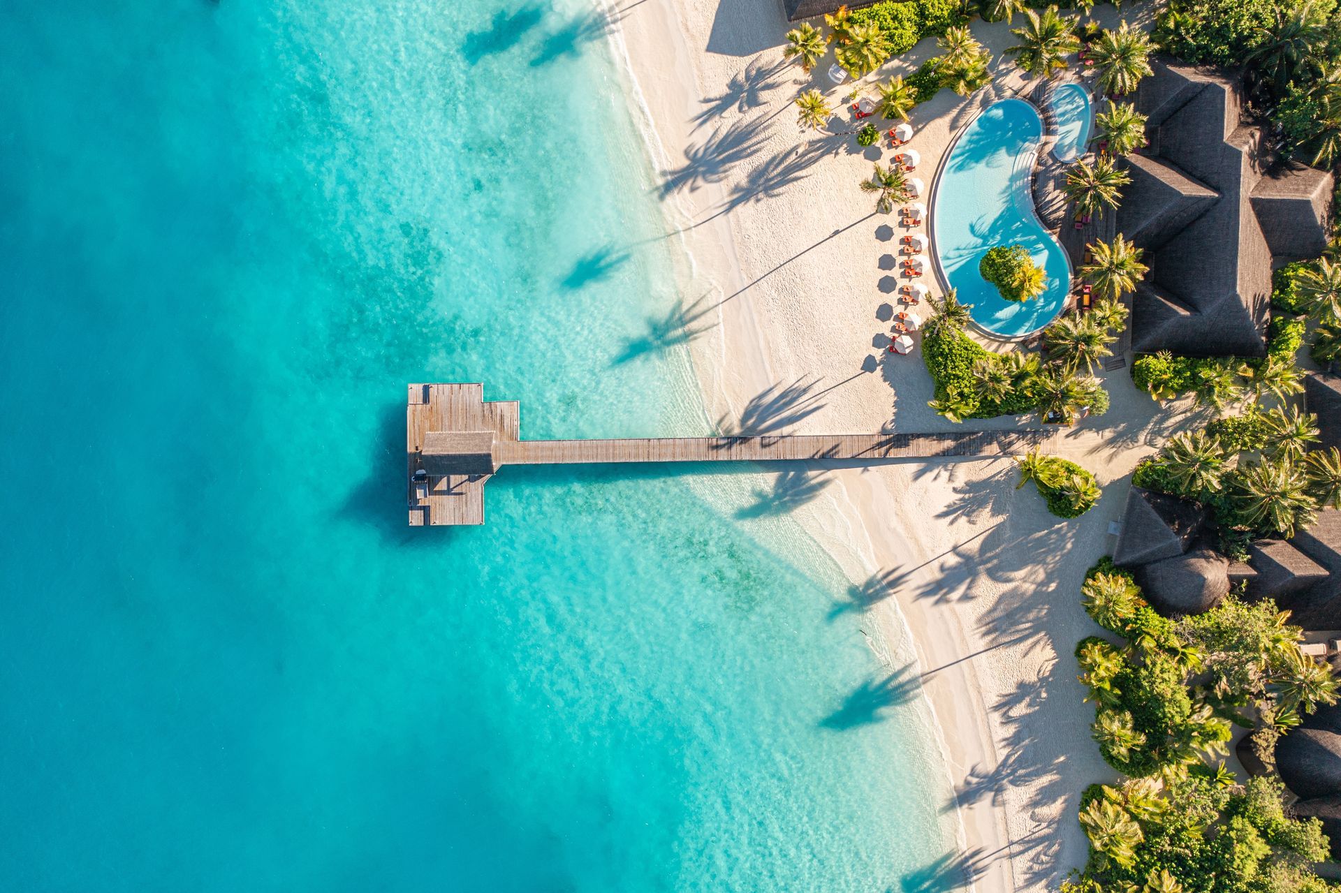 Aerial view of a tropical beach with turquoise water, white sand, a wooden pier, and resort poolside palms.