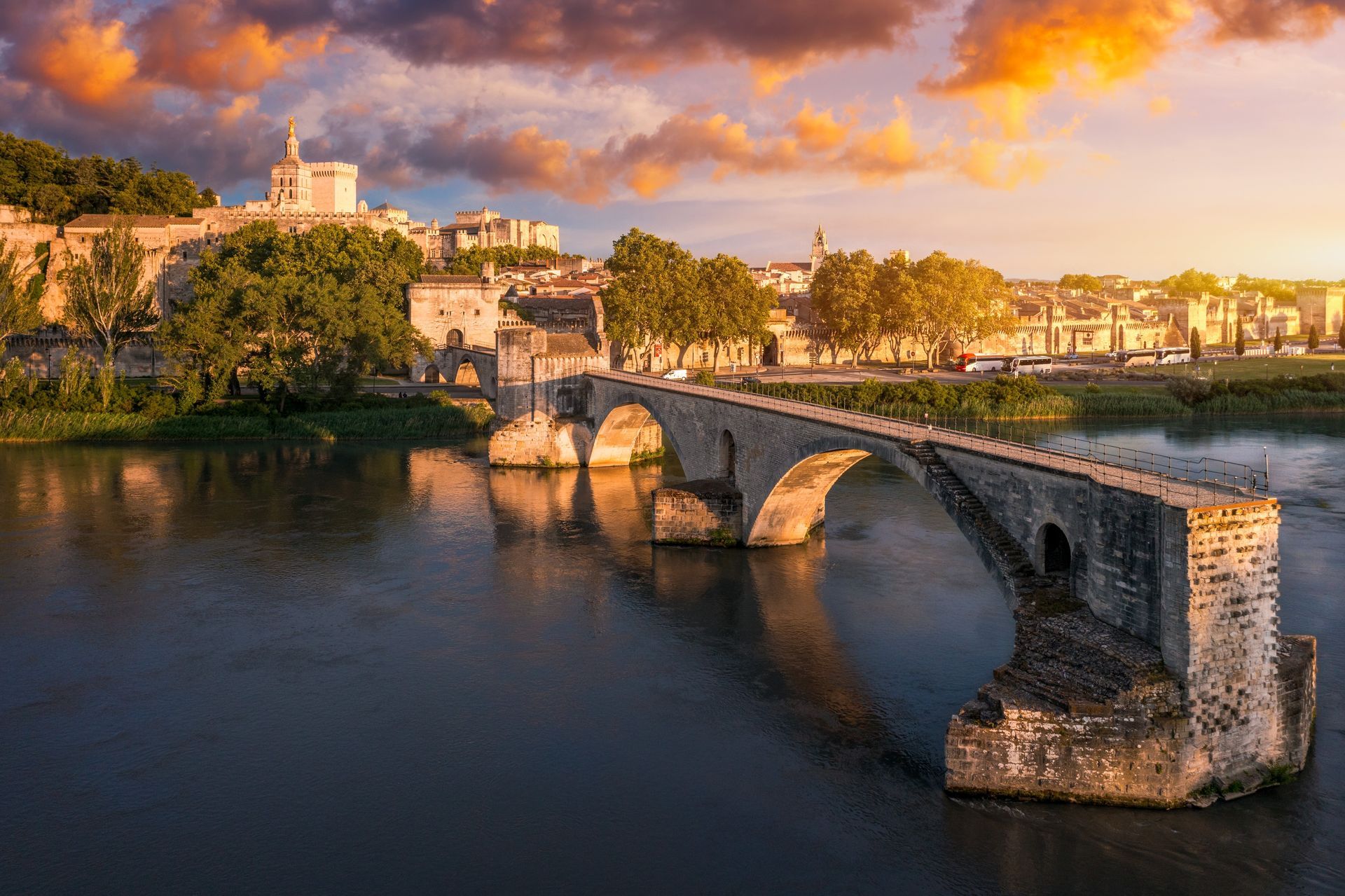 Stone bridge over a river at sunset, with warm clouds and historic buildings along the bank.