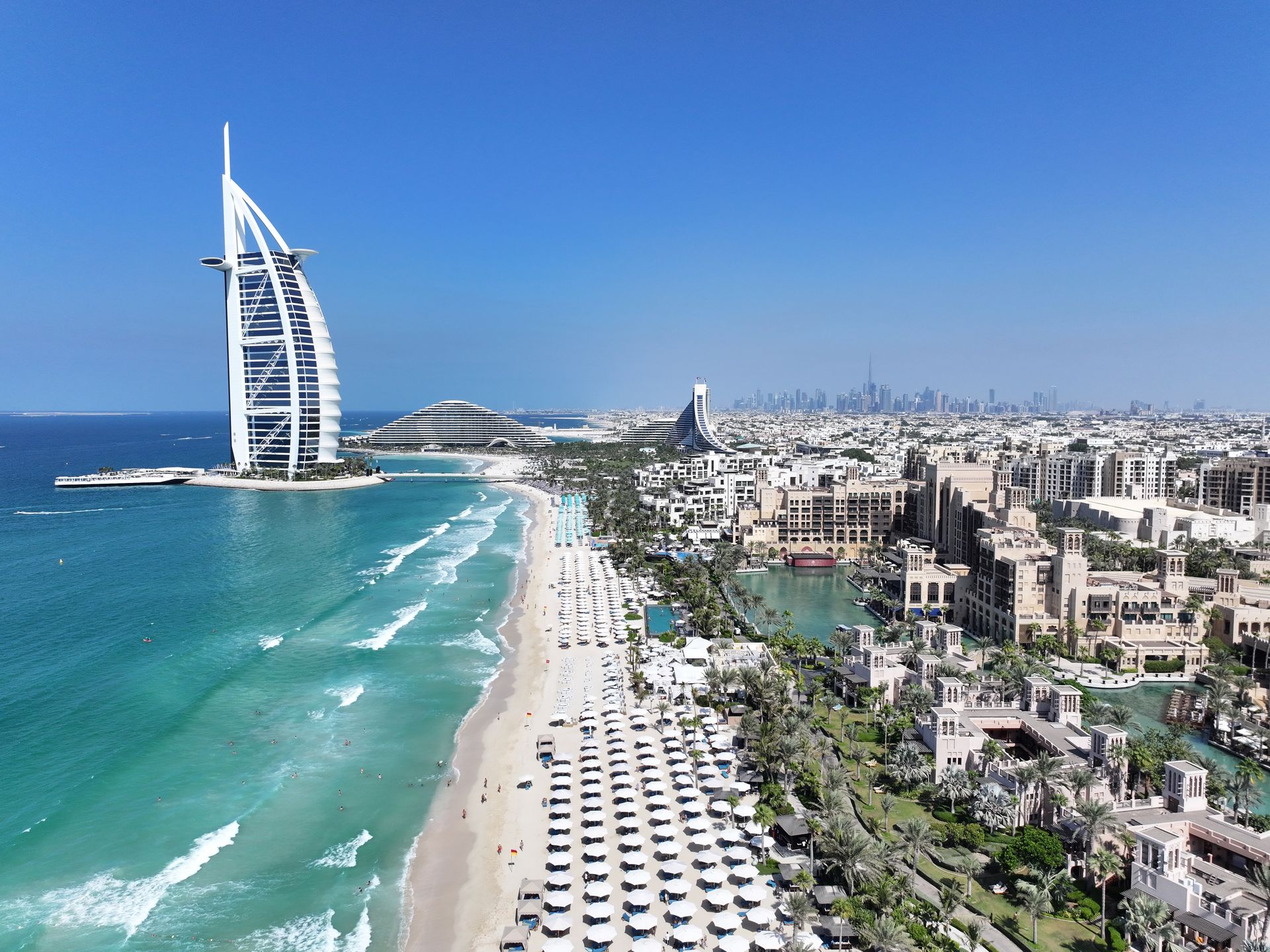 Aerial view of Dubai’s Burj Al Arab, beach, turquoise sea, and city skyline under a clear blue sky
