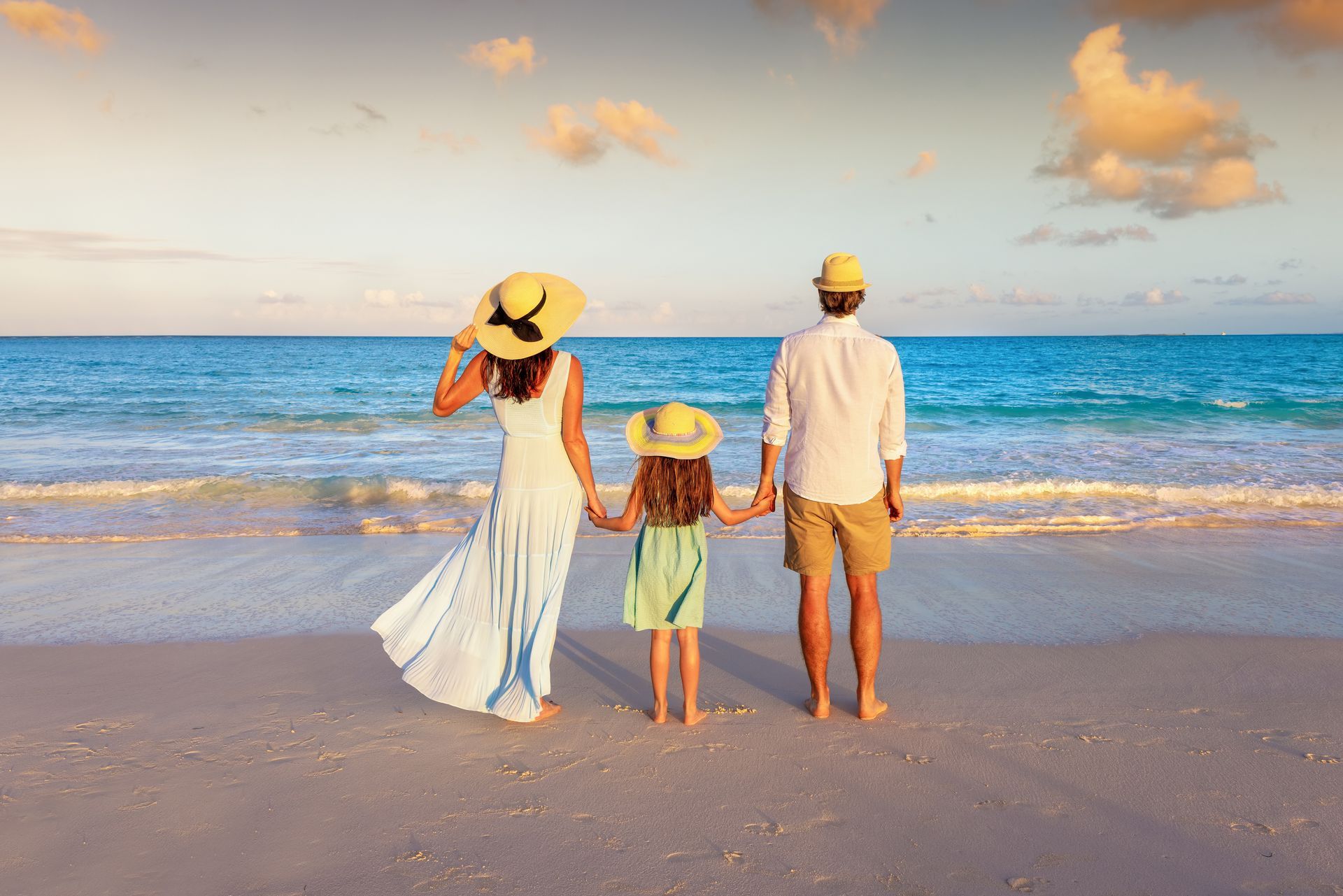 Three people in sun hats stand at the shoreline, facing the ocean at sunset.