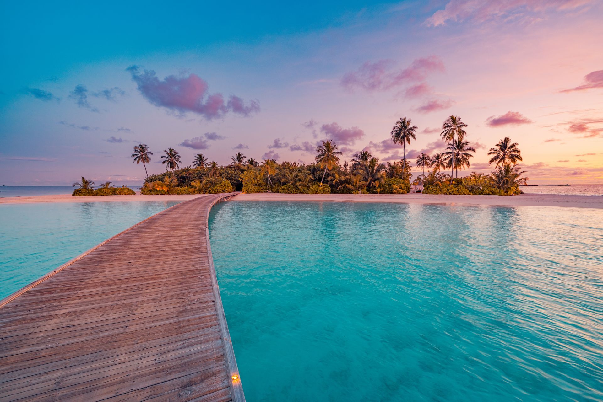 Wooden pier leading to a tropical island at sunset, with turquoise water and palm trees.