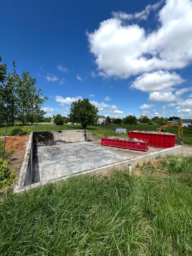 A rectangular concrete foundation sits in a grassy field under a sunny blue sky, with two large red dumpsters nearby.