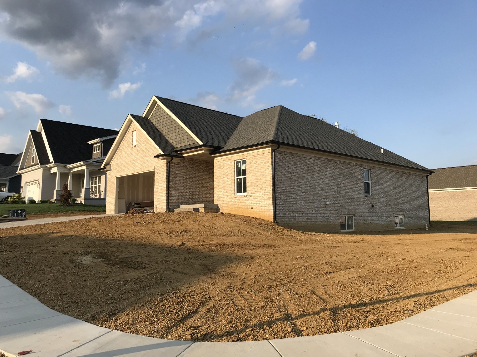 A newly constructed, light-brick ranch house with a dark roof and unfinished, dirt-covered yard on a suburban street.