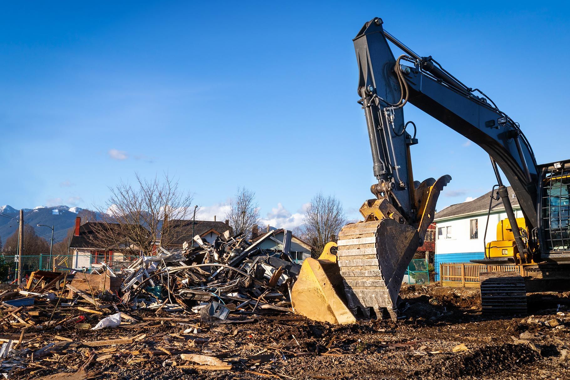 A large yellow excavator clears debris and piles of wreckage at a construction site against a clear blue sky.