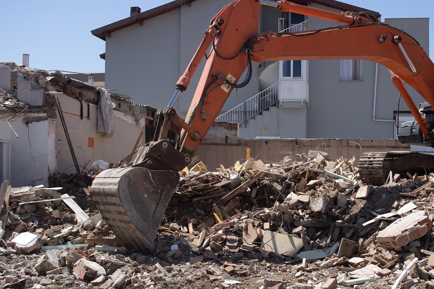 An orange excavator sits atop a pile of concrete rubble at a demolition site in front of a building.