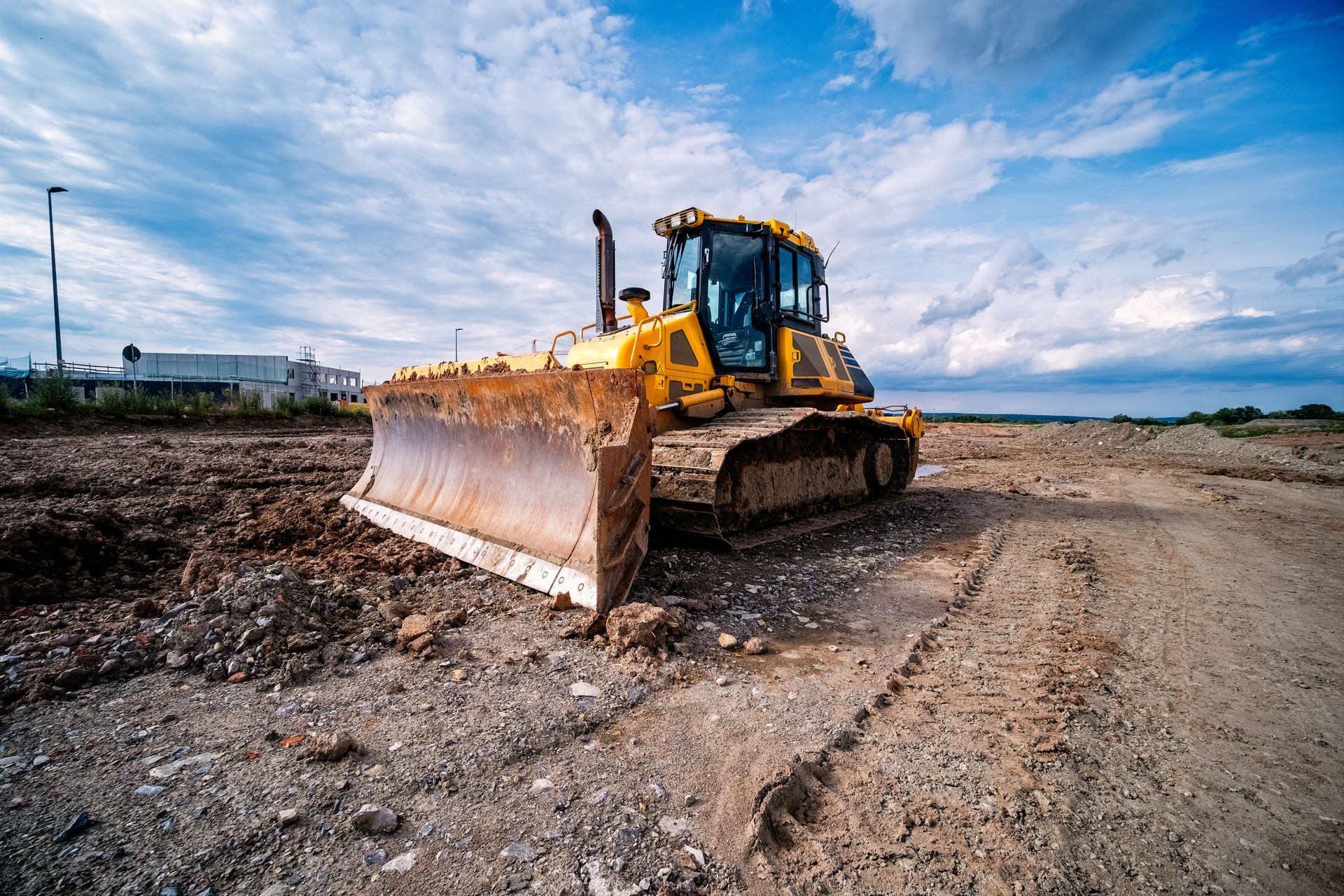 A yellow bulldozer sits on a rough, dirt construction site under a cloudy blue sky.