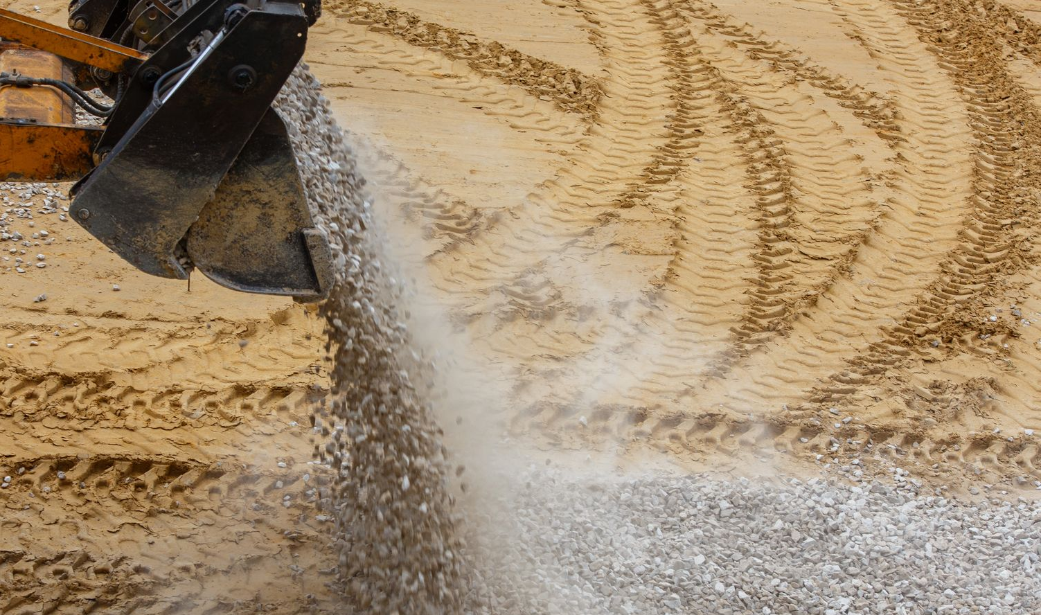 An excavator bucket pours gravel onto a sandy construction site with visible tire tracks.
