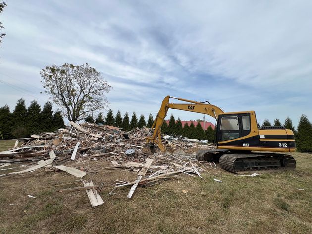 A yellow CAT excavator works to clear a large pile of debris from a demolished building in an open, grassy field.