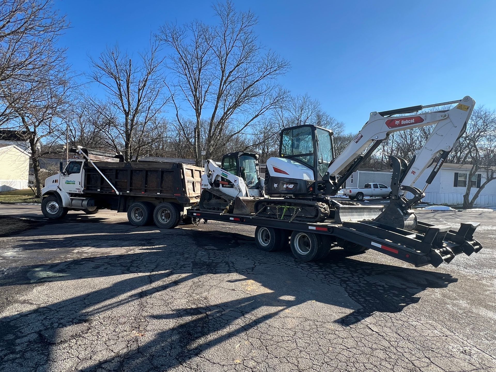 A white dump truck pulls a trailer carrying a white Bobcat excavator on a gravel lot under a clear blue sky.