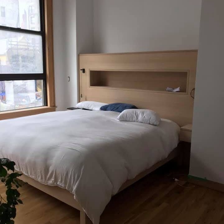 Minimal bedroom with a neatly made white bed, wood headboard, and a window on the left.
