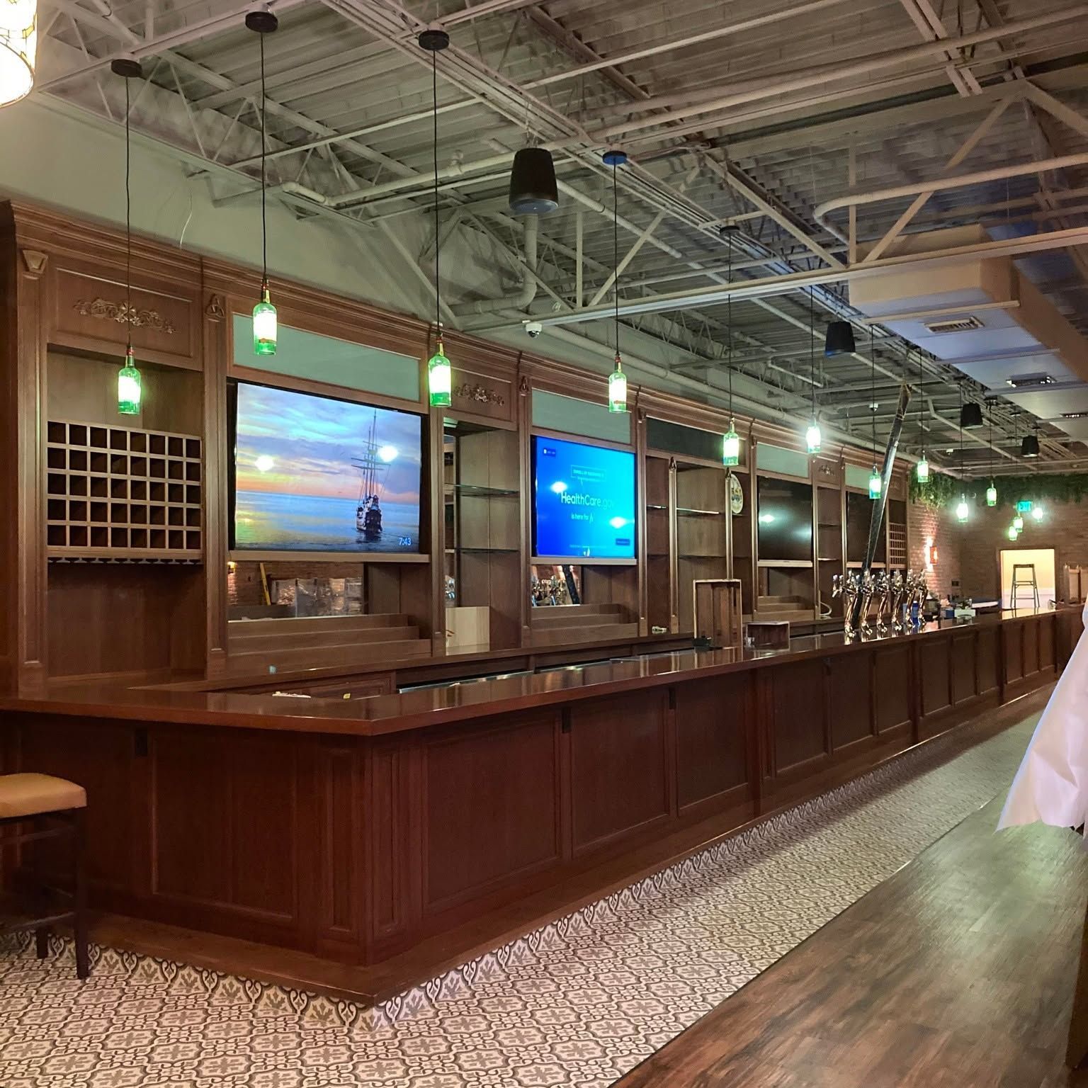 Empty bar interior with wood counter, tiled floor, and glowing green pendant lights under a high ceiling