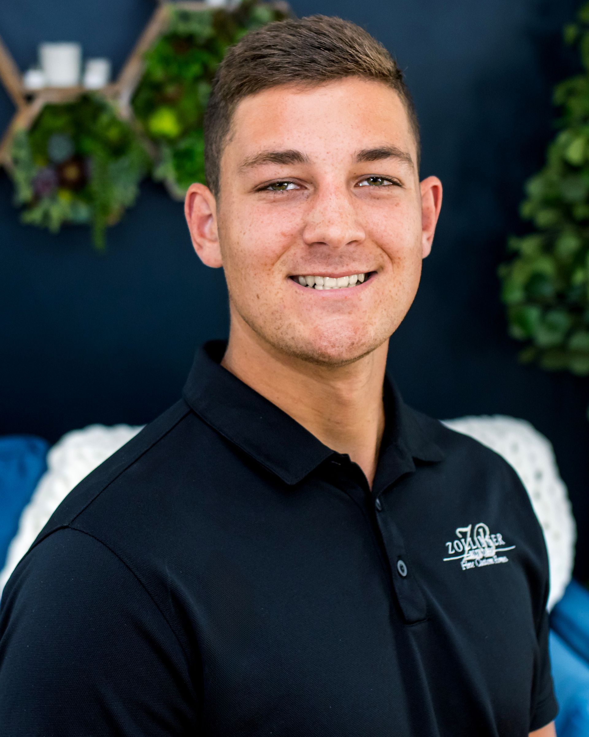 A person with short brown hair smiles while wearing a black polo shirt, positioned in front of a blue wall with greenery.